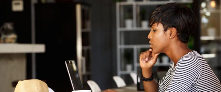 Photo of a woman concentrating on her laptop. She sits at a table and has a cup of coffee.